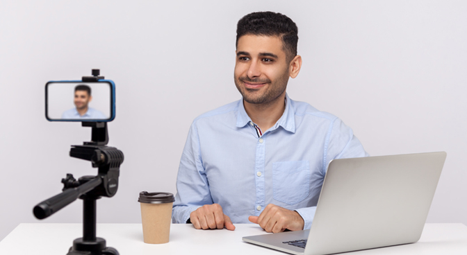 A teacher recording himself with his phone and a laptop is open near him.