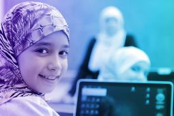 A hijab wearing student smiling while her laptop is open and her teacher in the background and is talking to the whole class.
