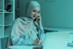 A woman wearing a hijab, and she is wring on her notebook while talking on her smartphone and her laptop is open in front of her.