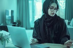 A woman wearing a hijab, checking her smartphone and using her laptop.
