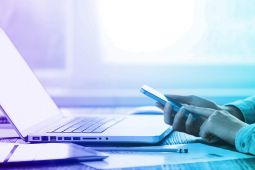 Hands of a female using a smartphone and in front of her an opened laptop.