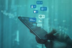 A hand holding a smartphone and symbols such as bubble speeches, likes and new messages are coming out of it.